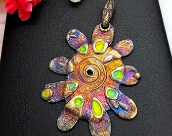 May include: A silver pendant with a colorful, abstract design. The pendant is shaped like a sun with a spiral in the center and green, yellow, and orange accents.