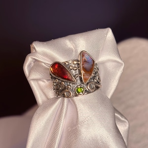 K&ouml;nnte beinhalten: Ein silberner Ring mit zwei gro&szlig;en, unregelm&auml;&szlig;ig geformten Edelsteinen. Einer ist tiefrot, der andere eine Mischung aus Orange und Blau. Zwischen ihnen befindet sich ein kleiner gr&uuml;ner Stein. Der Ring hat ein verziertes, strukturiertes Band.