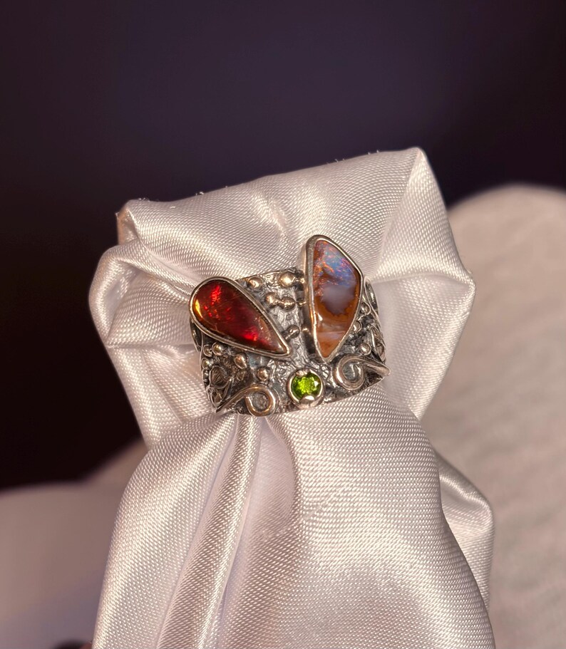 K&ouml;nnte beinhalten: Ein silberner Ring mit zwei gro&szlig;en, unregelm&auml;&szlig;ig geformten Edelsteinen. Einer ist tiefrot, der andere eine Mischung aus Orange und Blau. Zwischen ihnen befindet sich ein kleiner gr&uuml;ner Stein. Der Ring hat ein verziertes, strukturiertes Band.