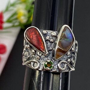 K&ouml;nnte beinhalten: Ein silberner Ring mit zwei gro&szlig;en, tropfenf&ouml;rmigen Steinen in Rot und Braun und einem kleinen gr&uuml;nen Stein. Der Ring hat ein verziertes, strukturiertes Design mit floralen und wirbelartigen Details.