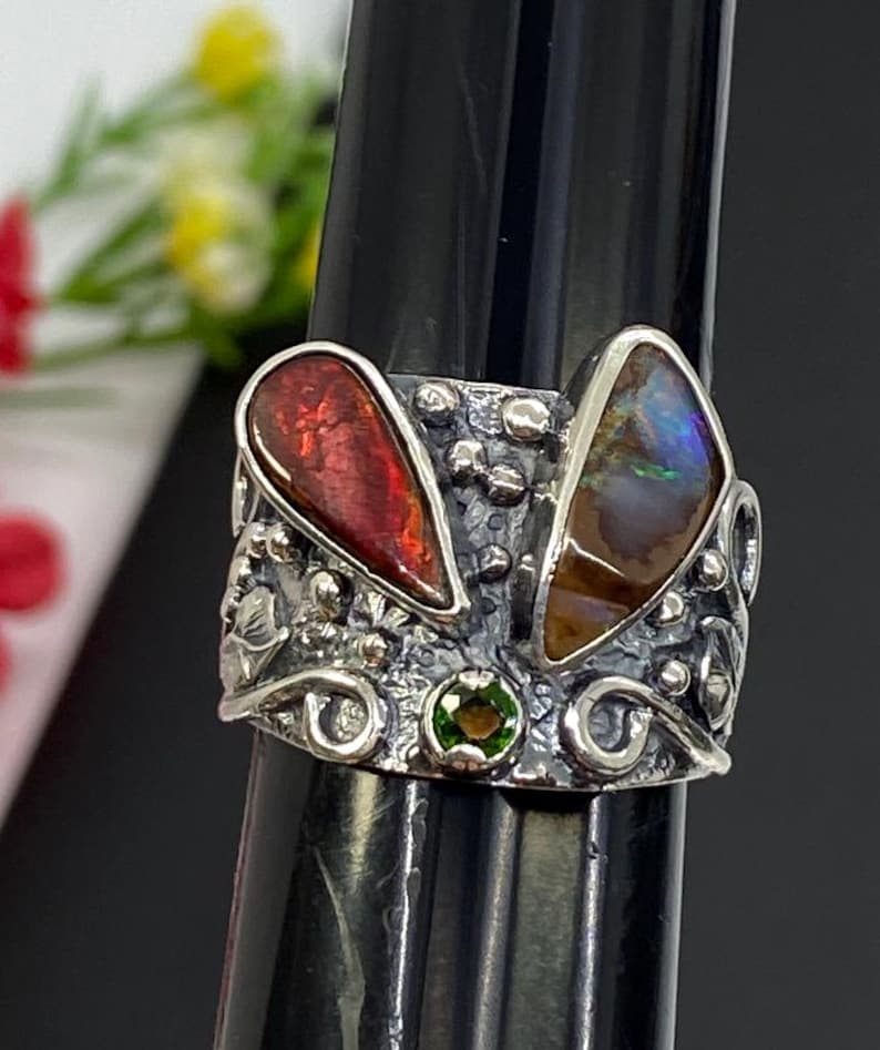 K&ouml;nnte beinhalten: Ein silberner Ring mit zwei gro&szlig;en, tropfenf&ouml;rmigen Steinen in Rot und Braun und einem kleinen gr&uuml;nen Stein. Der Ring hat ein verziertes, strukturiertes Design mit floralen und wirbelartigen Details.