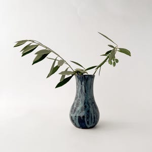 May include: A ceramic vase with a bulbous base and a narrow neck, featuring a glossy glaze in shades of deep navy blue and light grey with vertical drip patterns. A single olive branch with green leaves and small olives extends from the opening.