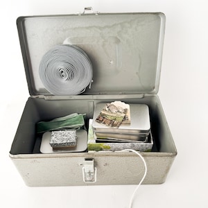 May include: A gray metal storage box with a hinged lid containing a roll of gray ribbon, a green velvet ribbon, a small rectangular box with a speckled gray lid, and a silver metal tin topped with a small rectangular paper tag.