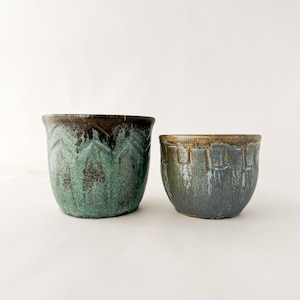 May include: Two ceramic planters with a rustic finish. The larger planter features a dark brown rim and a textured teal green body with a repeating chevron pattern. The smaller planter has a muted brown rim and a textured grey-blue body.