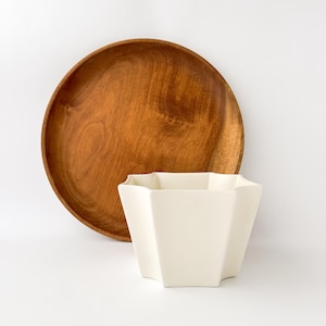 May include: A round, medium-brown wooden plate with a visible natural grain pattern positioned behind a white, six-sided ceramic planter or bowl with a smooth, matte finish and a flared, geometric rim design against a plain white background.