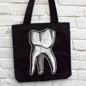 Op de afbeelding: Zwarte canvas tote bag met een witte afbeelding van een grote tand.