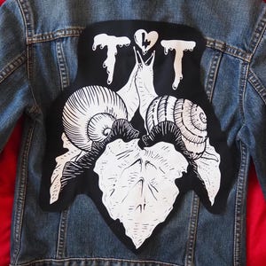 Könnte beinhalten: Blaue Jeansjacke mit großem schwarzem Aufnäher auf dem Rücken. Der Aufnäher zeigt eine weiße Illustration von Schnecken, Blättern und den Buchstaben "T" und einem Herzsymbol. Die Jacke hat Knopfverschlüsse und Taschen.