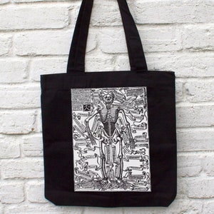 May include: Black tote bag featuring a white print of a skeleton with anatomical labels. The skeleton is standing with arms outstretched and legs slightly apart.