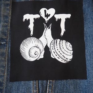 Könnte beinhalten: Schwarzer quadratischer Aufnäher mit einer weißen Grafik von zwei sich küssenden Schnecken, mit einem Herz und den Buchstaben "T T T" darüber. Der Aufnäher ist auf eine blaue Jeansjacke genäht.