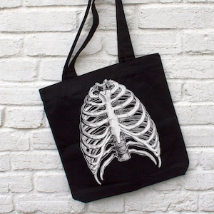Op de afbeelding: Zwarte tote bag met een witte anatomische illustratie van een ribbenkast.