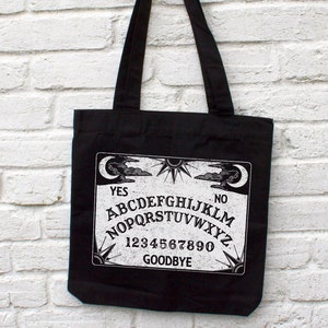 May include: Black canvas tote bag with a white graphic of a Ouija board. The board features the letters A-Z, numbers 0-9, and the words "YES", "NO", and "GOODBYE".