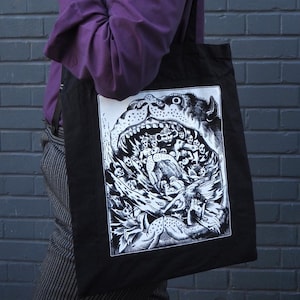 Peut inclure: Une personne portant une chemise violette et un chapeau noir porte un sac fourre-tout noir avec un imprimé graphique blanc d'une gueule de monstre avec de nombreuses figures à l'intérieur.