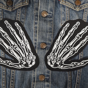 Op de afbeelding: Twee zwarte stoffen patches met witte skelethandjes op een blauw denim jack.