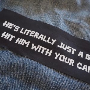 Op de afbeelding: Een zwarte patch genaaid op een blauw denim jack met witte tekst die "He's literally just a guy hit him with your car." luidt.
