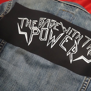 Op de afbeelding: Een zwart denim jack met een zwarte patch op de rug die "The Babe With The Power" zegt in witte gestileerde letters.
