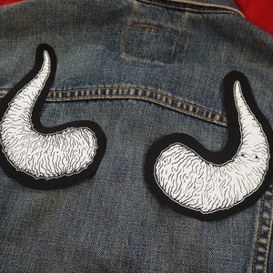 May include: Two black and white patches with a textured design of a curved horn shape. The patches are sewn onto a blue denim jacket.