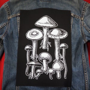 May include: A blue denim jacket with a black patch. The patch displays a detailed white illustration of several mushrooms of varying sizes and shapes. The jacket is placed on a red surface, providing a contrasting backdrop.