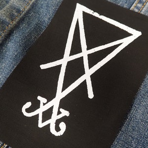 K&ouml;nnte beinhalten: Ein schwarzer, rechteckiger Aufn&auml;her mit einem wei&szlig;en Sigill-Design. Der Aufn&auml;her befindet sich auf einem blauen Denim-Stoff, wobei sich das wei&szlig;e Design vom dunklen Hintergrund abhebt. Wahrscheinlich f&uuml;r Kleidung oder Accessoires.