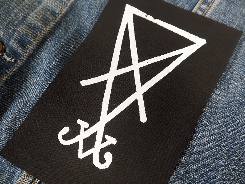 K&ouml;nnte beinhalten: Ein schwarzer, rechteckiger Aufn&auml;her mit einem wei&szlig;en Sigill-Design. Der Aufn&auml;her befindet sich auf einem blauen Denim-Stoff, wobei sich das wei&szlig;e Design vom dunklen Hintergrund abhebt. Wahrscheinlich f&uuml;r Kleidung oder Accessoires.
