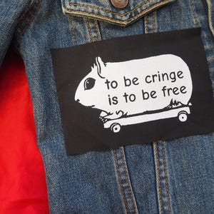 Könnte beinhalten: Jeansjacke mit einem schwarzen Aufnäher. Der Aufnäher zeigt ein weißes Meerschweinchen auf einem Skateboard mit dem Text "to be cringe is to be free". Die Jacke hat ein rotes Innenfutter.