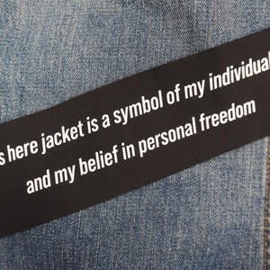 Peut inclure: Un patch rectangulaire noir avec du texte blanc qui dit : "...this here jacket is a symbol of my individuality, and my belief in personal freedom." Le patch est sur un fond en denim bleu.