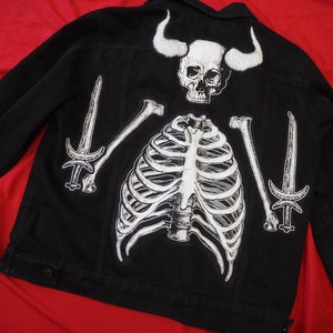 Op de afbeelding: Zwart denim jack met een wit skelet, ribbenkast en zwaarden ontwerp. Het skelet heeft hoorns en een schedel.