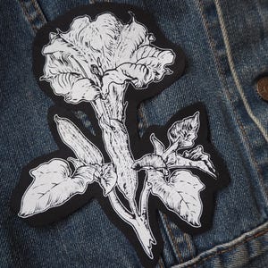 May include: A black fabric patch featuring a detailed white line art illustration of a Datura flower with its trumpet-shaped bloom, stem, and leaves. The design is rendered in a high-contrast style with intricate shading lines on the petals.