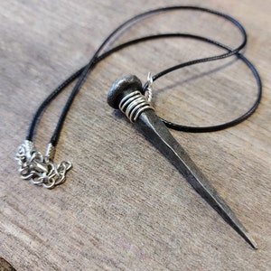 May include: A silver nail pendant necklace with a black cord. The nail is wrapped with silver wire at the top.