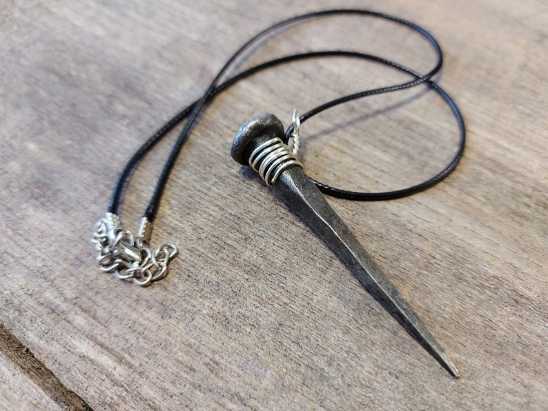May include: A silver nail pendant necklace with a black cord. The nail is wrapped with silver wire at the top.