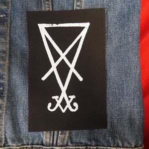 K&ouml;nnte beinhalten: Ein schwarzer Aufn&auml;her mit einem wei&szlig;en Sigill-Design, befestigt an einer Jeansjacke. Die Jacke hat messingfarbene Kn&ouml;pfe und einen roten Hintergrund.