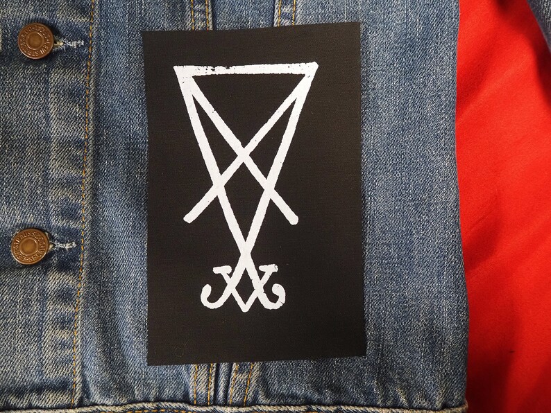 K&ouml;nnte beinhalten: Ein schwarzer Aufn&auml;her mit einem wei&szlig;en Sigill-Design, befestigt an einer Jeansjacke. Die Jacke hat messingfarbene Kn&ouml;pfe und einen roten Hintergrund.
