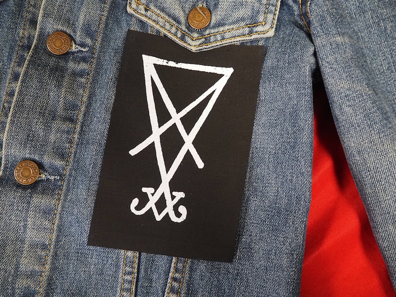 K&ouml;nnte beinhalten: Eine blaue Jeansjacke mit einem schwarzen Aufn&auml;her mit einem wei&szlig;en Sigill-Design. Die Jacke hat messingfarbene Kn&ouml;pfe und ein rotes Futter, das in der offenen Front sichtbar ist.