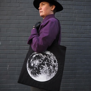 Może przedstawiać: Osoba w fioletowym swetrze i czarnym kapeluszu trzyma czarną torbę na zakupy z białym nadrukiem księżyca.