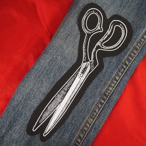 以下が含まれることがあります： デニムジャケットに、白黒のハサミのパッチが施されています。パッチは、白いアウトラインでハサミの詳細なイラストを特徴としています。背景は鮮やかな赤い生地です。