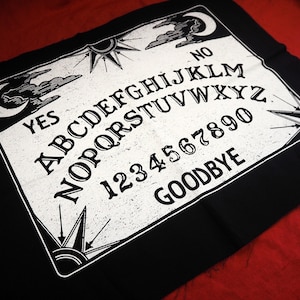 Può includere: Tessuto nero con un design di tavola Ouija stampato in bianco. La tavola presenta le lettere A-Z, i numeri 1-9 e 0, le parole "YES", "NO" e "GOODBYE".