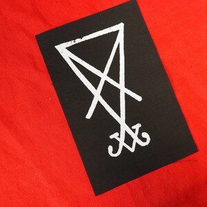 K&ouml;nnte beinhalten: Ein schwarzer rechteckiger Aufn&auml;her mit einem wei&szlig;en Sigill-Design. Das Sigill ist ein stilisiertes Symbol mit sich kreuzenden Linien und einem Dreieck oben. Der Aufn&auml;her befindet sich auf einem leuchtend roten Hintergrund.