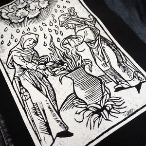 May include: Black and white patch featuring a woodcut-style illustration of two figures tending a vase with flames. Rain falls from a stylized sun above. The patch is rectangular and set against a denim jacket.