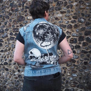 May include: A light blue denim vest with a large black and white graphic on the back featuring a skull, mushroom, and snail design. The vest is worn over a black shirt and paired with dark grey jeans.