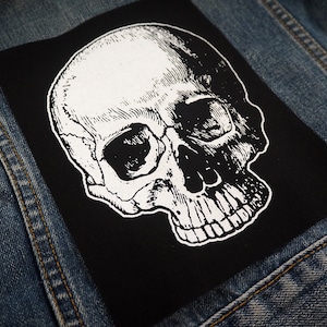 May include: A black and white skull patch with a detailed illustration of a human skull. The patch is iron-on and can be used to decorate clothing, bags, or other items.