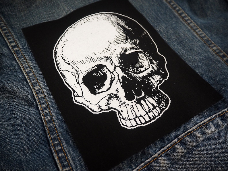 May include: A black and white skull patch with a detailed illustration of a human skull. The patch is iron-on and can be used to decorate clothing, bags, or other items.