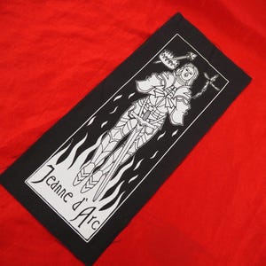 Op de afbeelding: Zwart-witte patch met een gestileerde afbeelding van een vrouw in harnas die een zwaard vasthoudt. De tekst "Jeanne d'Arc" is gedrukt in een wit banner aan de onderkant van de patch.