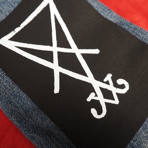 K&ouml;nnte beinhalten: Ein schwarzer Aufn&auml;her mit wei&szlig;en Symbolen, auf eine Jeansjacke gen&auml;ht. Der Aufn&auml;her hat ein wei&szlig;es, umgekehrtes Dreieck und andere Symbole. Der Jeansstoff ist blau. Der Hintergrund ist rot.