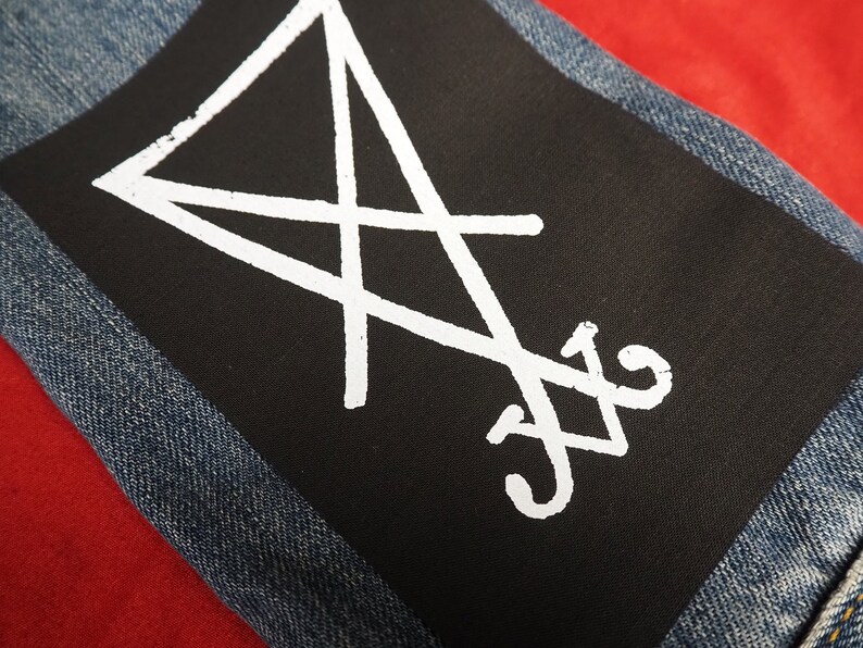 K&ouml;nnte beinhalten: Ein schwarzer Aufn&auml;her mit wei&szlig;en Symbolen, auf eine Jeansjacke gen&auml;ht. Der Aufn&auml;her hat ein wei&szlig;es, umgekehrtes Dreieck und andere Symbole. Der Jeansstoff ist blau. Der Hintergrund ist rot.