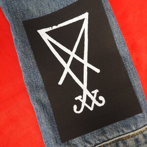 K&ouml;nnte beinhalten: &Auml;rmel einer Jeansjacke in Blau mit einem schwarzen Aufn&auml;her. Der Aufn&auml;her zeigt ein wei&szlig;es, stilisiertes Symbol aus Linien und Kurven. Das Symbol befindet sich auf schwarzem Hintergrund. Der Hintergrund ist rot.
