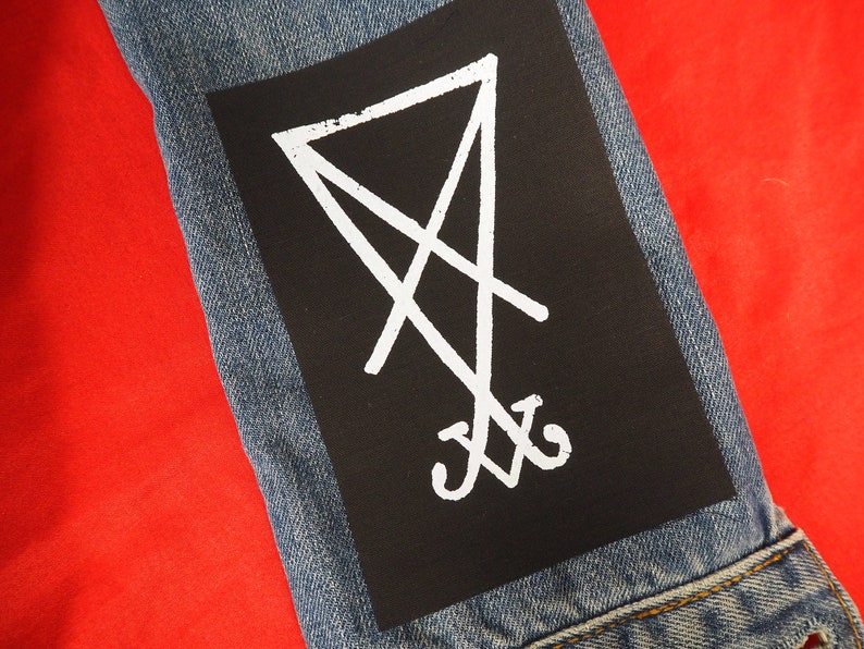 K&ouml;nnte beinhalten: &Auml;rmel einer Jeansjacke in Blau mit einem schwarzen Aufn&auml;her. Der Aufn&auml;her zeigt ein wei&szlig;es, stilisiertes Symbol aus Linien und Kurven. Das Symbol befindet sich auf schwarzem Hintergrund. Der Hintergrund ist rot.
