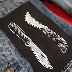 Peut inclure: Deux illustrations en noir et blanc de couteaux sur un patch de tissu noir. Les couteaux sont de style vintage.