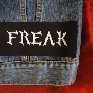 May include: A black rectangular patch with the word "FREAK" in white gothic lettering is affixed to a denim jacket. The jacket is blue and has visible stitching and a button. The background is a red fabric.