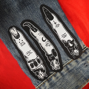 May include: Three black and white iron-on patches depicting dripping, skeletal fingers with different nail designs. The patches are sewn onto a blue denim jacket.