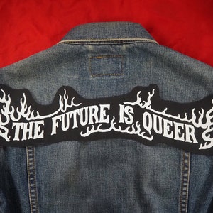 May include: A denim jacket with a black patch on the back that says "THE FUTURE IS QUEER" in white letters with flames on either side.