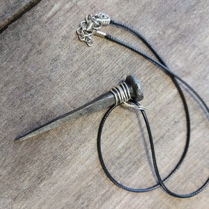 May include: A black cord necklace with a large, silver, nail-shaped pendant. The nail is wrapped with wire near the top.